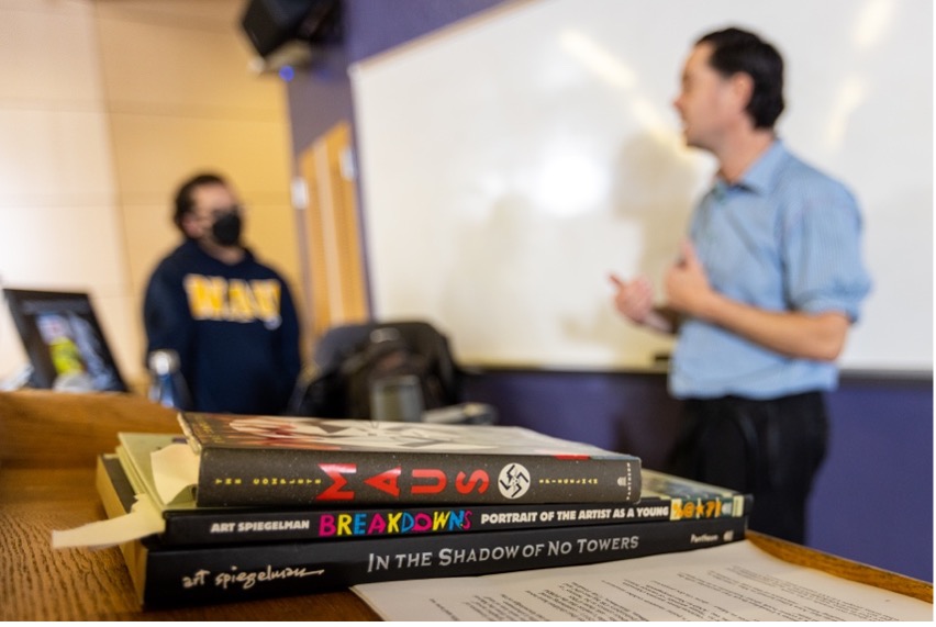 Pile of books in foreground with professor and student in background discussing books.