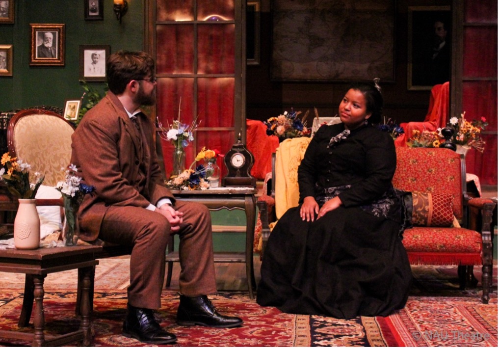 Two students in period dress on stage with victorian set pieces.