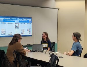 Two women and one man sit in front of a digital monitor showing an intern poster.