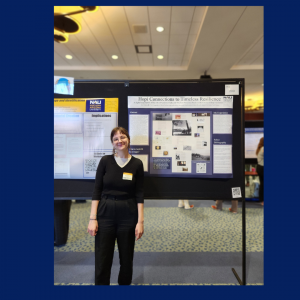 A woman stands in front of a poster wearing all black and a pair of glasses.