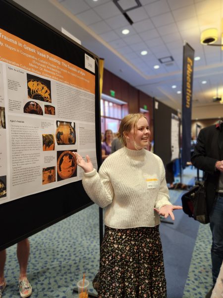 A woman in a white sweater and flowered skirt gestures toward a orange and white poster. She has blonde hair pulled back.