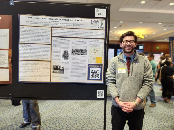 A tall man with dark hair and beard with glasses poses with a blue and yellow poster. 