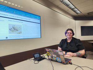 A student with short hair and glasses sites in front of a monitor.
