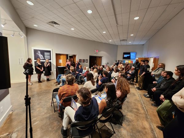 Image depicts a group of people seated in a gallery facing 3 student speakers. In the background are works of art.