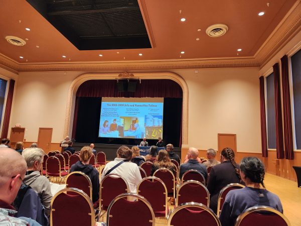 The image shows a panel of speakers at the front of a room with an audience facing away from the camera.