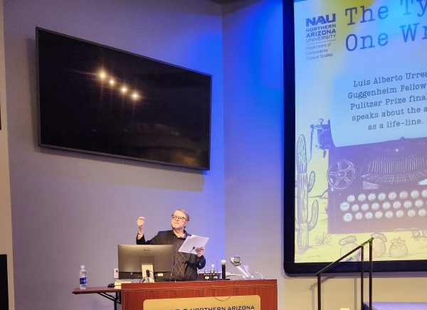Author Luis Urrea stands behind a podium with one hand raised. He wears glasses and a black jacket and shirt.