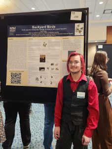 A man in a maroon shirt and black vest with glasses stands in front of a research poster.