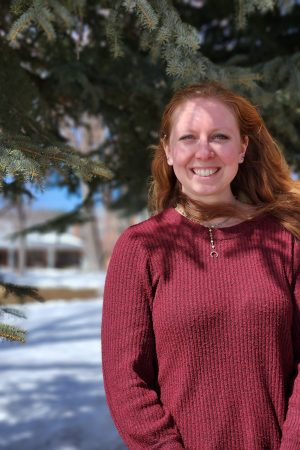 The image shows a woman with long red hair against a snowy background and a pine tree wearing a red sweater. 