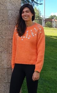 Woman with dark hair and orange sweater stands against a cement wall.