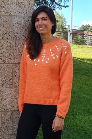 Woman with dark hair and orange sweater stands against a cement wall.