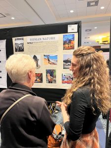 A woman with long hair shares her work with an older woman with white hair in front of a beige research poster.