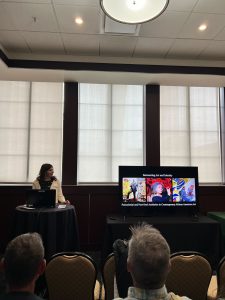 A woman with dark hair stands at a podium next to a monitor with images of art. 