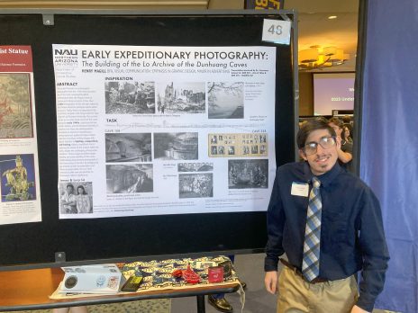 student in tie and glasses smiles in front of a poster