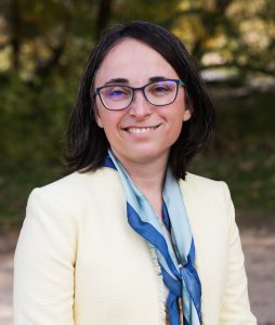 The image shows a woman with dark hair wearing glasses, a white suit jacket and a blue scarve,