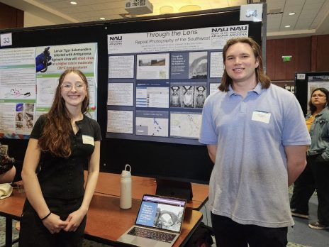 two students smiling in front of a poster and laptop