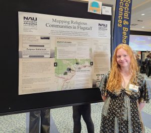 A woman with long red hair stands in front of a green, blue and beige research poster.