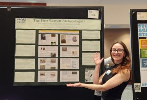 A woman wearing glasses stands in front of a green research poster.