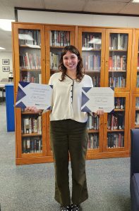 A woman with dark hair in a white blouse stands holding two certificates in front of a bookcase. 