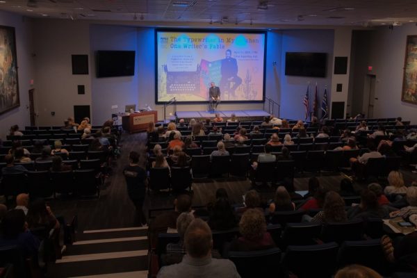 Luis Urrea sits on a stool on a stage in front of full assembly hall.