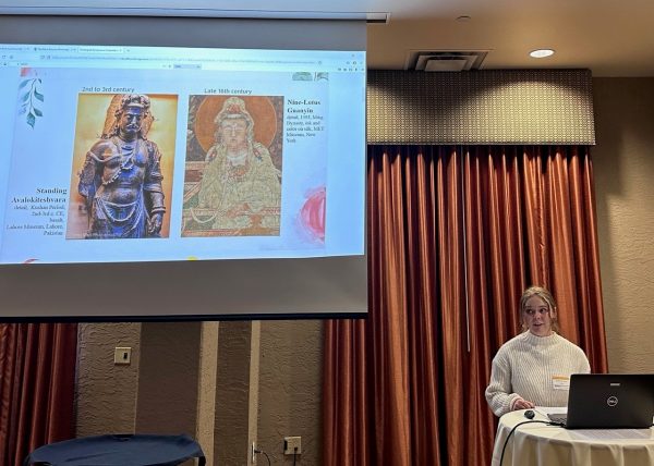A blond woman in a white sweater stands in front of a podium with a red curtain behind her and with a computer in front of her. On the screen behind her is images of the Buddha from medieval China.