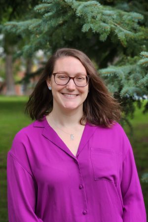 The image shows a woman with brown hair and glasses wearing a pink shirt against a background of pine trees.