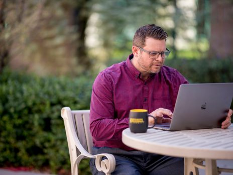 man in red shirt sitting at a table outdoors using a laptop