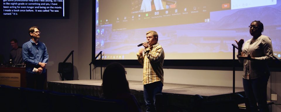 3 people presenting on a stage with the image of 3 others show on a screen behind them