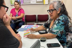 A woman showing a colorful card to a man at a desk while another woman looks on.