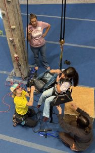 girl in a rock climbing gym being lifted in an accessible climbing chair by a lady as another lady watches