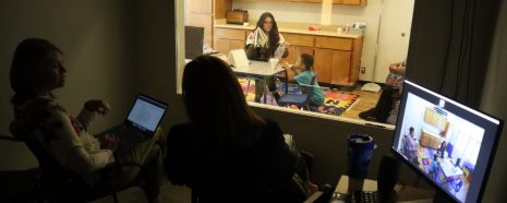 2 people in a dark observation room watching through a window as a woman works with a small child at a table