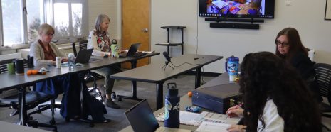 four women sit working with papers and computers with a TV screen in the background displaying an observation room