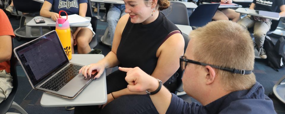 Justice Armfield is sitting with a student and gesturing towards the screen on her laptop. 
