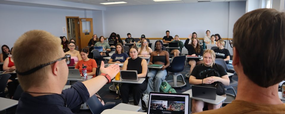 2 presenters in front of a class of students. One is gesturing towards a student who's hand is raised.