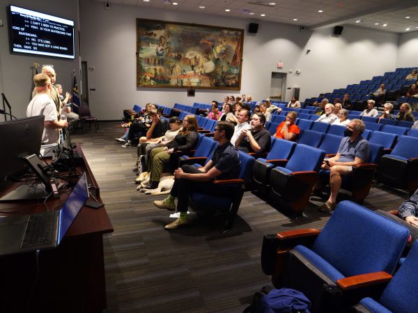 People seated in a theater watching people presenting in front of a screen showing small text.