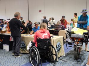 People talking around tables in an exhibit hall there is a man in a wheelchair in the foreground and another man pushing a one wheeled chair made for hiking