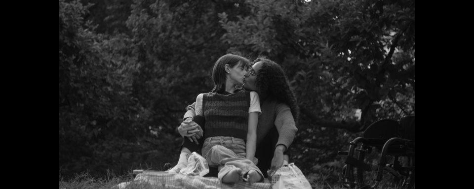 2 people sitting on a blanket outside kissing with trees and a wheelchair in the background