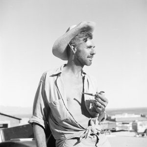 [Tad Nichols with straw hat and corncob pipe - Part of Cliff Dwellers Lodge in background; this image appears in Tad's Glen Canyon book on the title page.]