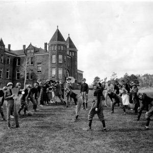 [Girls' High Jinks: Group of young women dressed in football uniforms and as men along with two men, in front of the Old Main-Ashurst buildings, Northern Arizona Normal School]