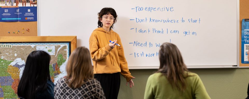 A CUPI Participant in a classroom outlining obstacles to higher education on a whiteboard, facing a panel of three women in front of them.