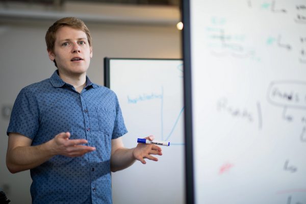 Joseph Mihaljevic is standing next to a white dry erase board with a blue dry erase marker in his left hand. He is talking and holding both hands up, and is wearing a blue short sleeve collared button-down shirt with spots in a pattern.