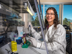Emily Cope smiles in her lab. She is wearing a white lab coat and is wearing glasses and latex gloves and holding a dropper and a test tube.