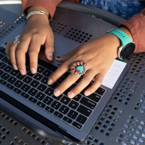 Close-up of hands with turquoise jewelry typing on a laptop keyboard at a metal outdoor table.