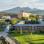 Aerial view of a university campus with modern buildings and green space, with large mountains in the background.