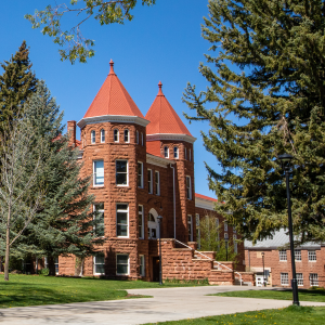 Historic red sandstone building with turrets and red roofs, framed by pine trees on a green lawn under a blue sky.