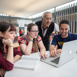 An instructor stands over three college students collaborating around a laptop and an open textbook.