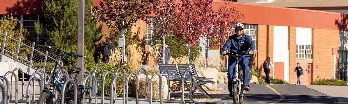Outdoor urban scene with a large yellow 'NAU' sign, a person riding a bicycle, bike racks, and mountainous landscape in the background.