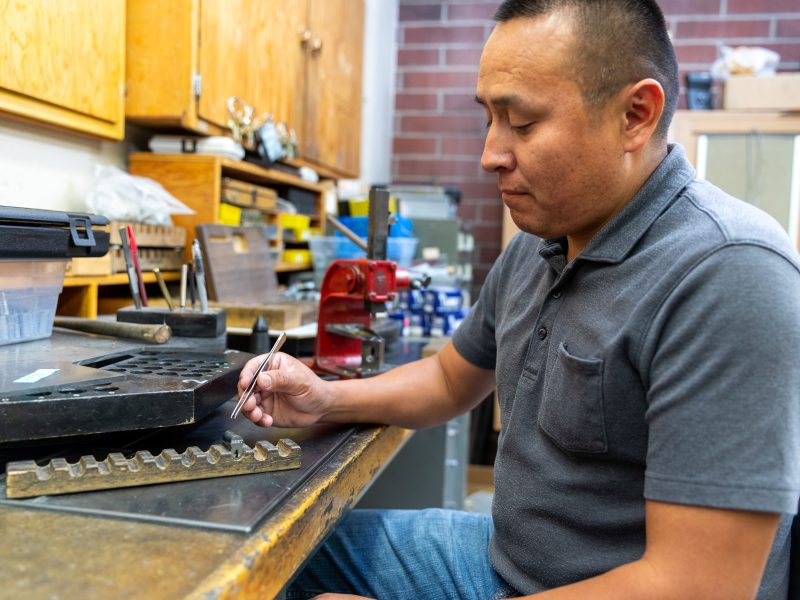 A person is sitting at a workbench in a workshop setting, focused on a task involving tools and equipment.