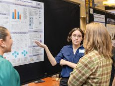 Several individuals standing in front of a display board with charts, diagrams, and text. The display board appears to have information about a research or academic project. The individuals are engaged in discussion or presentation of the content on the display board.