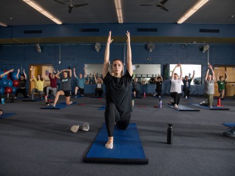College female in a yoga class on mat in yoga pose with arms straight up in the air, bending one leg.