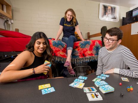 three college students in a dorm room, 2 women and 1 man, playing a board game